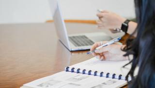 An academic writing in a notebook and using a laptop at a desk